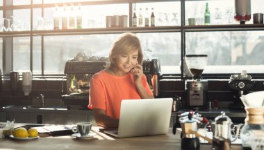 Dizüstü bilgisayarda çalışan ve smartphone üzerinde konuşurken orta yaşlı bir kadın barista