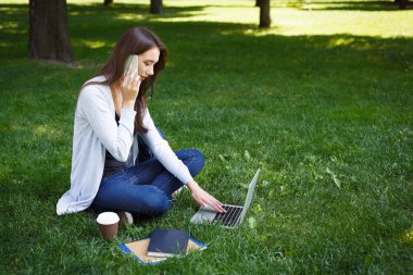 Laptop ile çalışma ve parkta telefonda konuşurken genç esmer kadın bir çimenlerin üzerinde