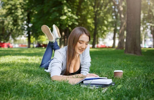 Joven mujer asiática tomando notas, acostada sobre una hierba verde. Sonriendo y tomando café en el parque durante el día —  Fotos de Stock