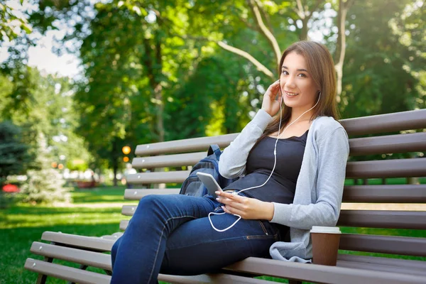Jovem estudante de TI sorridente em fones de ouvido. Ouvir música e beber café no parque — Fotografia de Stock