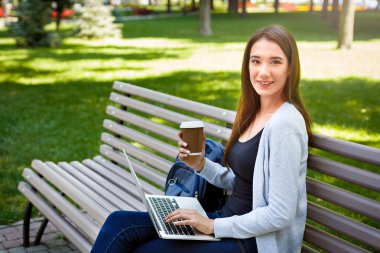 Parkta motive serbest meslek. Bir bankta, kahve içme ve laptop ile çalışma gülümseyen kız