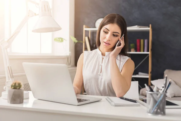 Ung kvinna som arbetar på laptop och consulting på telefon — Stockfoto