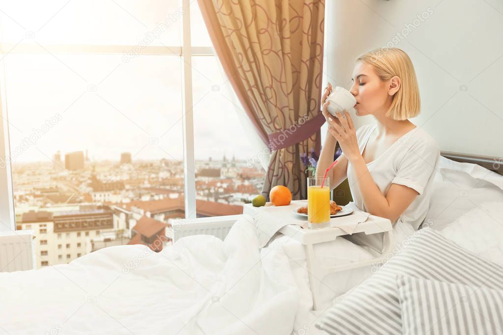 Beautiful young woman sitting on bed and having breakfast
