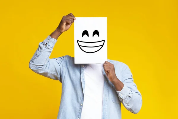 African man hiding face behind paper with drawn happy emoticon — Stock Photo, Image