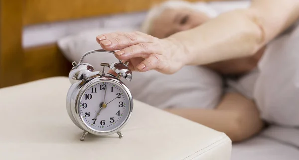 Ανώτερη γυναίκα γυρίζοντας το ξυπνητήρι, ξαπλωμένη στο κρεβάτι, πανόραμα — Φωτογραφία Αρχείου