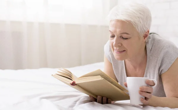 Спокойная пожилая женщина лежит на кровати с книгой и кофейной чашкой — стоковое фото
