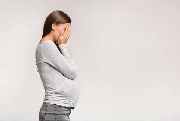 Depresyjna kobieta w ciąży zakrywająca twarz stojąca na szarym tle — Zdjęcie stockowe