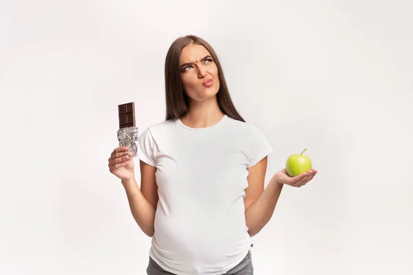 Έγκυος κορίτσι επιλέγοντας μεταξύ μήλου και σοκολάτας στέκεται, Studio Shot — Φωτογραφία Αρχείου