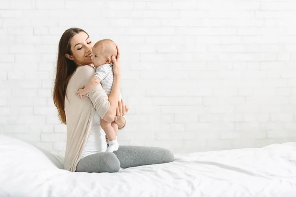 Young woman embracing adorable baby sitting on bed at home — Stock Photo, Image