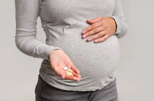 Chica embarazada irreconocible mostrando píldoras de pie sobre fondo gris, recortado —  Fotos de Stock