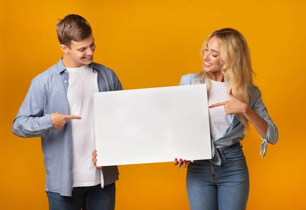 Millennial ζευγάρι κρατώντας κενό πίνακα διαφήμισης και δείχνοντας σε αυτό — Φωτογραφία Αρχείου