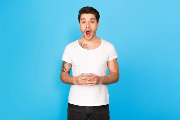 Shock content. Surprised guy holding smartphone, blue background — Stock Photo, Image