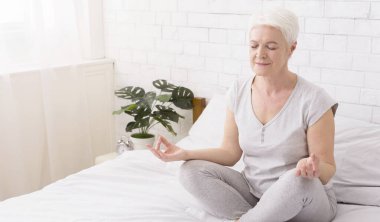 Yaşlı kadın yoga yapıyor, evde yatağında lotus pozisyonunda meditasyon yapıyor.