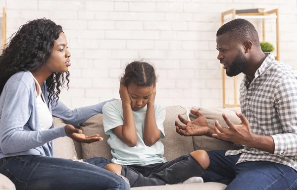 Masentunut pieni musta tyttö istuu välillä väittelevät vanhemmat, sulkemalla korvat — kuvapankkivalokuva