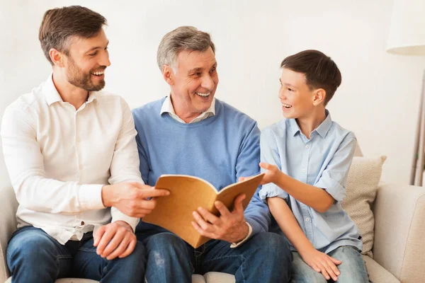 Hijo, padre y abuelo leyendo libro sentado en el sofá interior —  Fotos de Stock