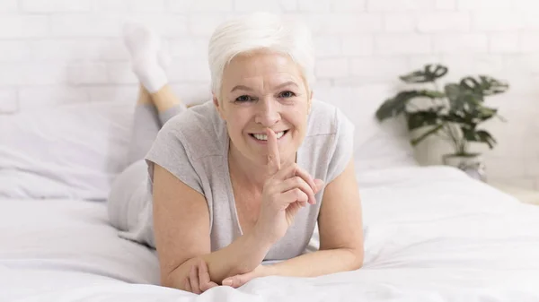 Misteriosa donna anziana che tiene il dito sulla bocca, sdraiata a letto — Foto Stock