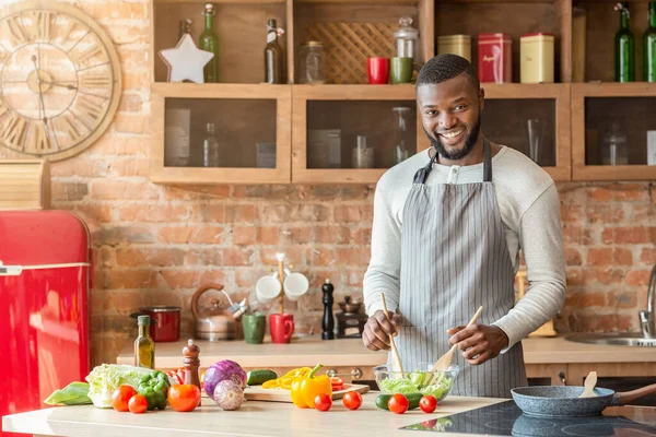 Sorrindo homem negro preparando salada saudável fresca na cozinha — Fotografia de Stock