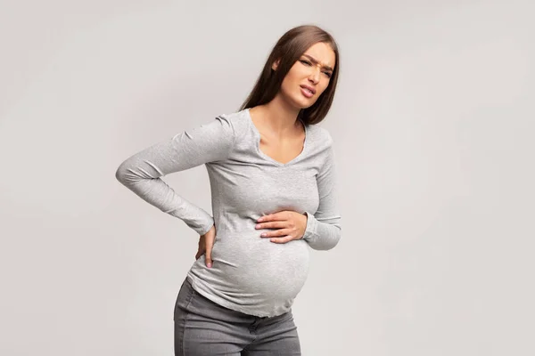 Gravid dam lider av ryggsmärta stående över grå bakgrund — Stockfoto