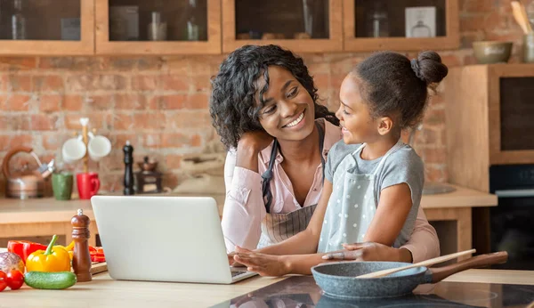 Mamá y su hija abrazando, la lectura de la receta en el ordenador portátil en la cocina — Foto de Stock