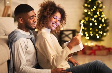Heyecanlı Afro-Amerikalı çift Noel ağacının önünde oturma odasında Noel hediyesini açıyor.