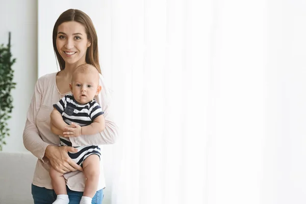 Bella donna che tiene in braccio il neonato, in piedi vicino alla finestra — Foto Stock