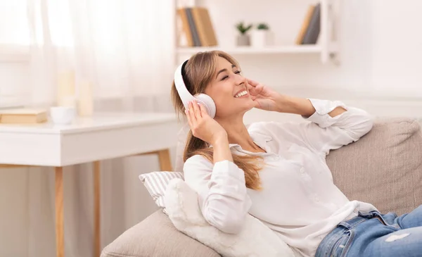 Dama con auriculares escuchando música tumbada en el sofá en casa —  Fotos de Stock