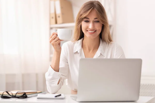 Χαμογελαστή επιχειρηματίας κρατώντας φλιτζάνι καφέ κάθεται στο φορητό υπολογιστή στο γραφείο — Φωτογραφία Αρχείου