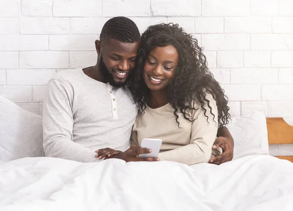 Sorrindo casal preto usando smartphone sentado na cama juntos — Fotografia de Stock