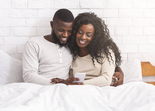 Smiling black couple using smartphone sitting on bed together