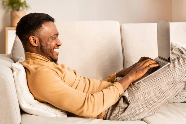 Gelukkig Afro-Amerikaanse man met behulp van laptop liggen op bank Indoor — Stockfoto