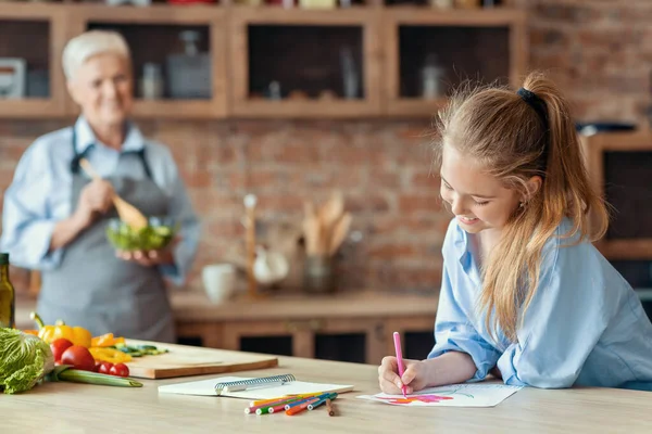 Kleines Mädchen zeichnet in der Küche, während Oma hilft — Stockfoto
