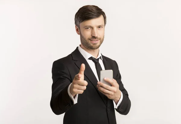Empresário segurando telefone apontando o dedo para a câmera de pé, fundo branco — Fotografia de Stock