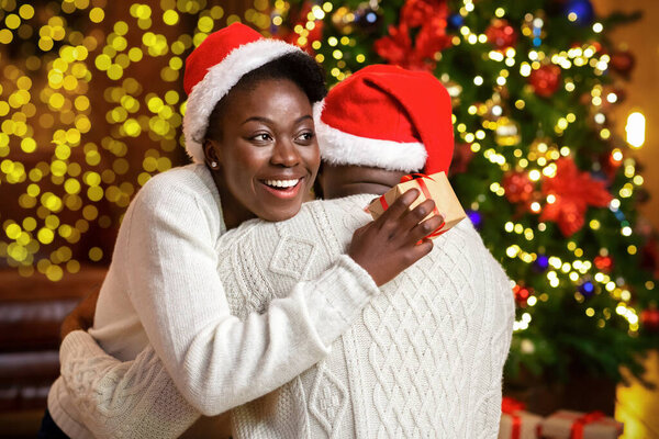 Благодарная чёрная жена держит рождественский подарок и обнимает мужа
