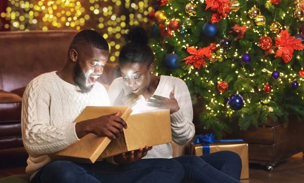 Чорна пара, що відкриває різдвяну подарункову коробку у вітальні біля ялинки — стокове фото