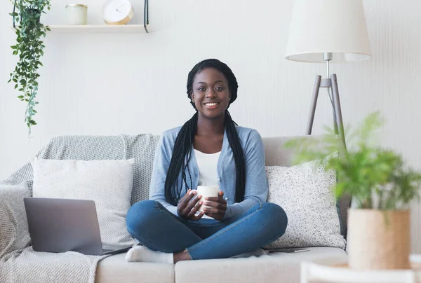 Donna afro freelance seduta sul divano con caffè e laptop — Foto Stock