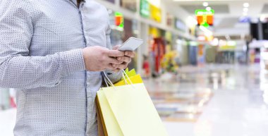 Cep telefonuyla yeni kıyafetler arayan, alışveriş merkezinde yürüyen bir adam.