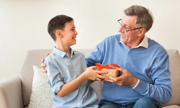 Boy Giving Grandfather Birthday Gift Sitting On Sofa Indoor