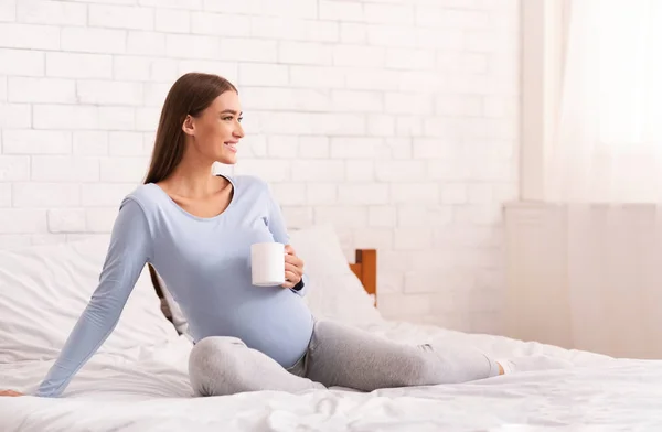 Mulher grávida feliz bebendo café sentado na cama no quarto — Fotografia de Stock