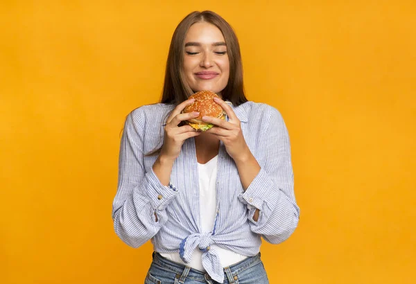 Mujer alegre oliendo hamburguesa de pie sobre fondo amarillo — Foto de Stock