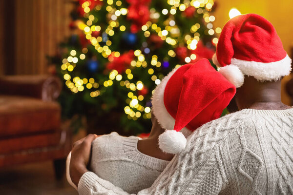 Black couple in Santa hats sitting next to Christmas tree
