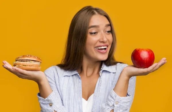 Menina com fome em pé escolhendo entre maçã e hambúrguer, fundo amarelo — Fotografia de Stock