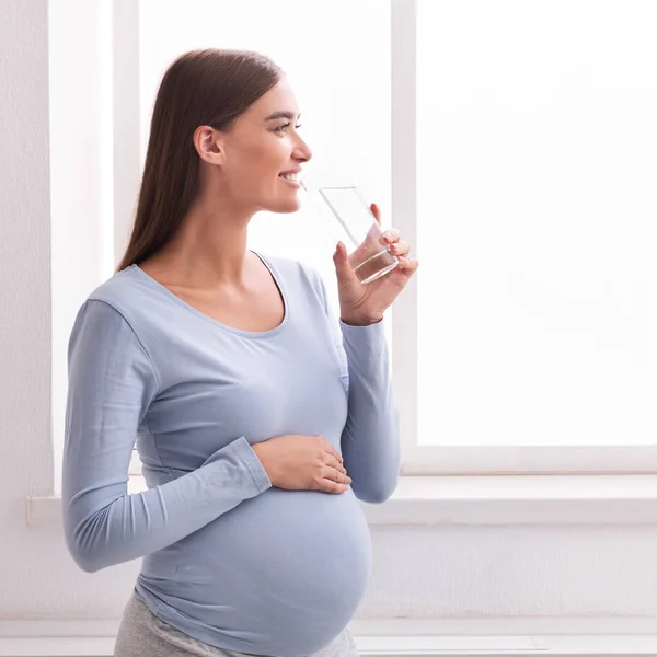 Positief zwanger meisje drinken water staande op zoek in het venster Indoor — Stockfoto