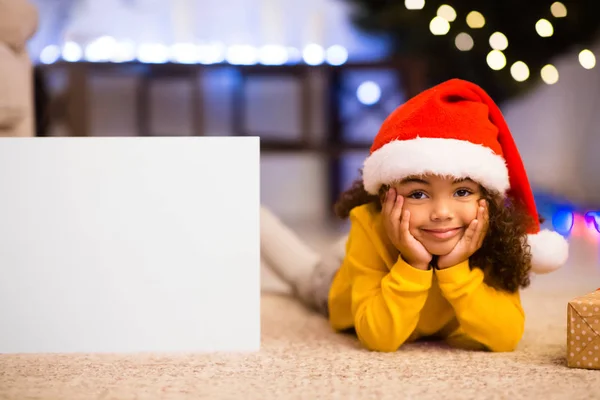 Симпатична маленька африканська дівчинка в капелюсі Санта лежить поруч з порожнім плакатом — стокове фото