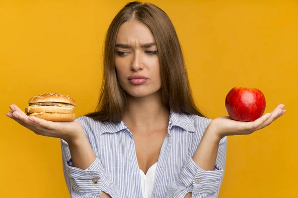 Lady Holding Burger And Apple Standing Choosing Food, Studio Shot Royalty Free Stock Images