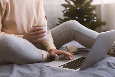 Kadın yatakta oturuyor, sıcak kakao içiyor ve dizüstü bilgisayar kullanıyor.