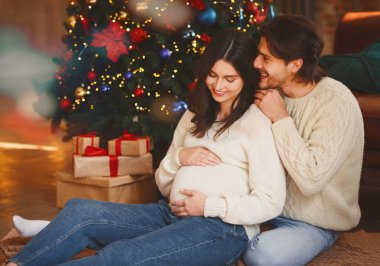 Hamile kadın ve kocası Noel ağacının yanında oturuyor.