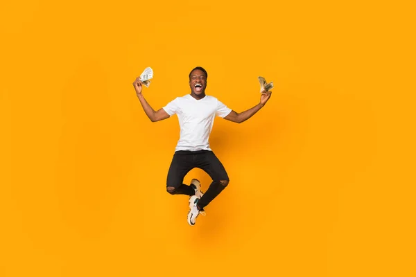 Emocionado hombre afro sosteniendo el dinero saltando en el aire, fondo naranja —  Fotos de Stock