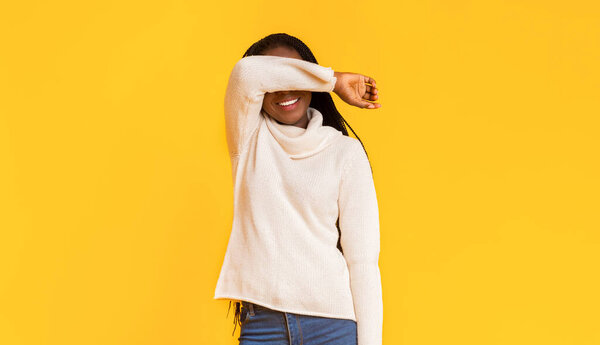 Black shy winter girl in sweater covering her face with arm, hiding from someone, yellow background, copy space