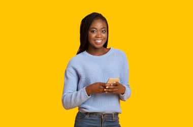 Gülümseyen siyah kadın sarı arka planda akıllı telefon kullanıyor.
