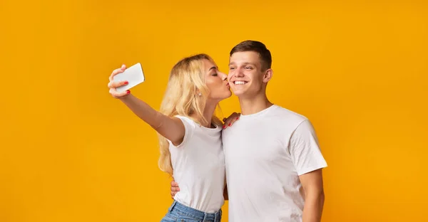 Chica joven tomando selfie mientras besa a su novio en la mejilla — Foto de Stock